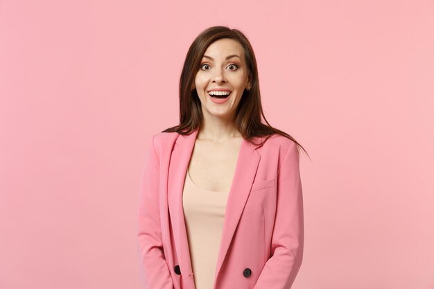 Portrait of excited smiling young woman wearing jacket standing and looking camera isolated on pastel pink wall background in studio. People sincere emotions lifestyle concept. Mock up copy space.