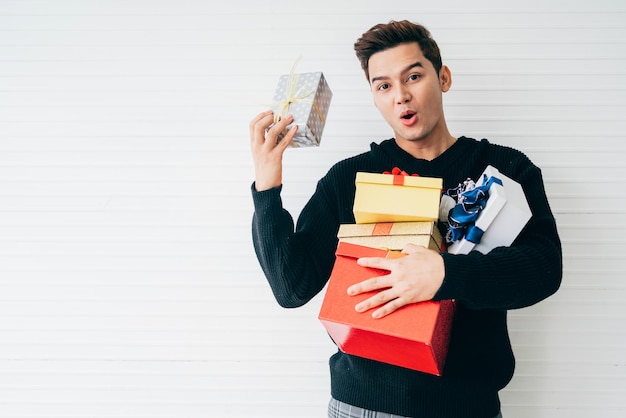 Photo portrait of an excited and surprised young asian man wearing christmas sweater holding many gifts