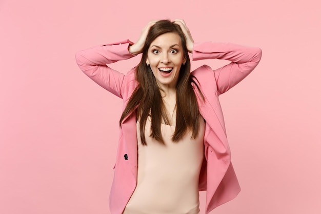 Portrait of excited surprised young woman in jacket looking camera, putting hands on head isolated on pastel pink background in studio. People sincere emotions, lifestyle concept. Mock up copy space.