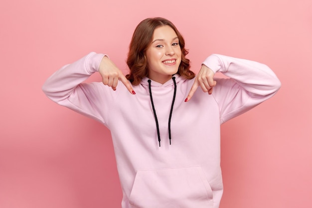 Portrait of excited teen curly haired female in hoodie pointing down place for commercial idea, looking at camera with toothy smile. Indoor studio shot isolated on pink background