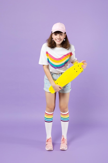 Portrait of excited teen girl in vivid clothes standing, holding yellow skateboard isolated on violet pastel wall background in studio. People sincere emotions, lifestyle concept. Mock up copy space.