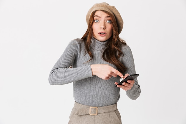 Portrait of excited woman 30s wearing hat smiling and holding cell phone isolated on white