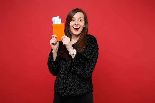 Portrait of excited woman in black fur sweater holding passport, boarding pass ticket isolated on bright red wall background in studio. People sincere emotions, lifestyle concept. Mock up copy space.