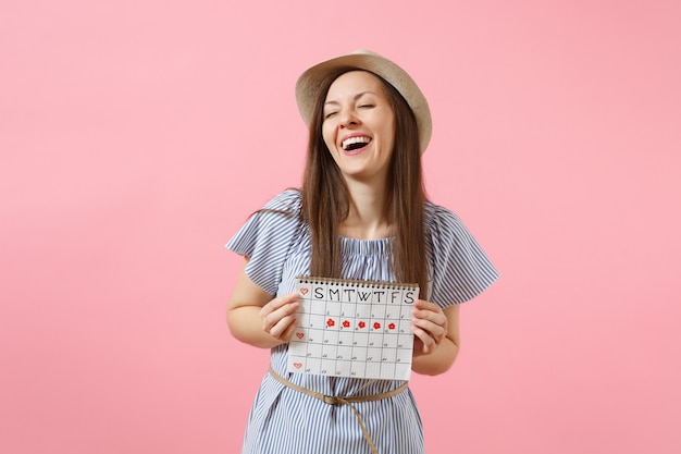 Portrait of excited woman in blue dress, hat holding periods calendar for checking menstruation days isolated on bright trending pink background. Medical, healthcare, gynecological concept. Copy space