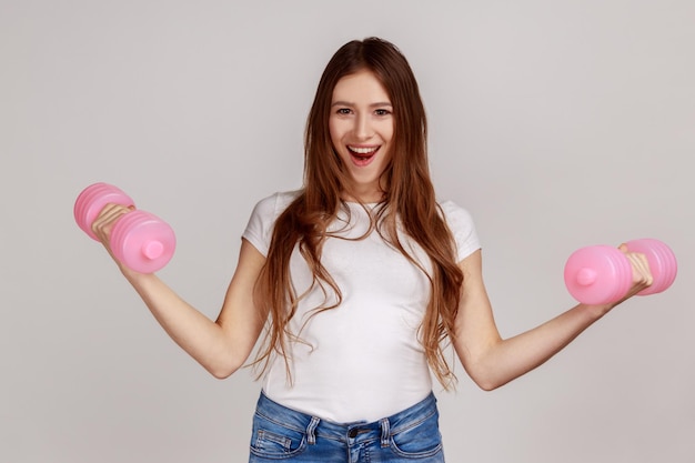 Photo portrait of excited woman standing with raised arms and holding pink dumbbells active lifestyle wearing white casual style tshirt indoor studio shot isolated on gray background