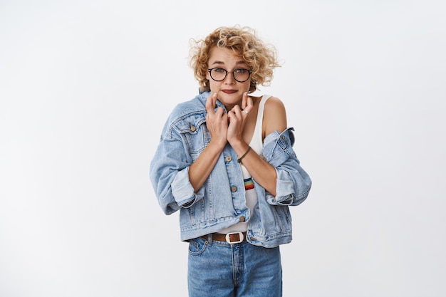 Portrait of excited worried and impatient cute stylish blond girl with glasses in denim jacket smirking nervously cross fingers for good luck as praying for dream fulfilled