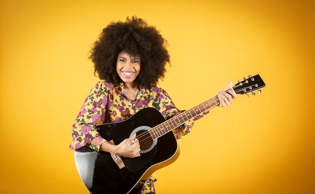 Portrait of excited young african american woman with a bright\
smile dressed in casual clothing dancing with a guitar over yellow\
background