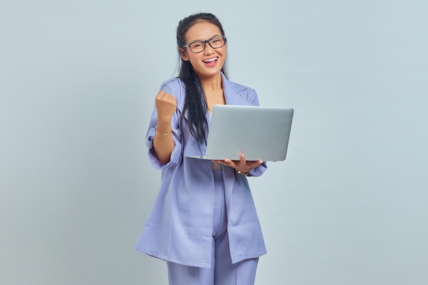 Портрет взволнованной молодой азиатской женщины, стоящей с ноутбуком и празднующей удачу в новой работе на белом фоне
