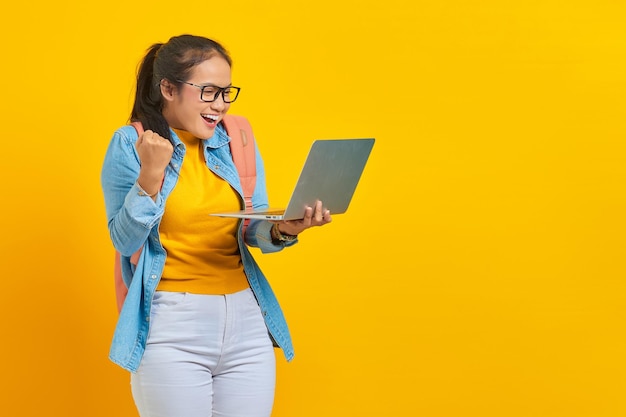 Portrait of excited young asian woman student in casual clothes with backpack using laptop and celebrating success isolated on yellow background education in university college concept