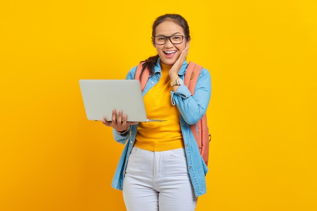 Portrait of excited young asian woman student in casual clothes with backpack using laptop and touching cheek with hands isolated on yellow background education in college university concept