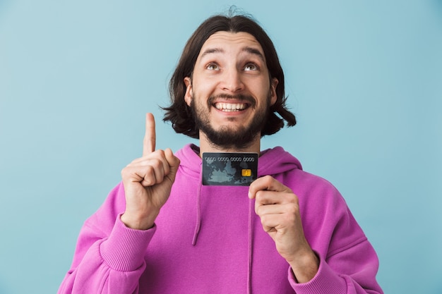 Портрет возбужденного молодого бородатого мужчины в повседневной одежде, стоящего изолированно над стеной, показывая кредитную карту, указывая вверх