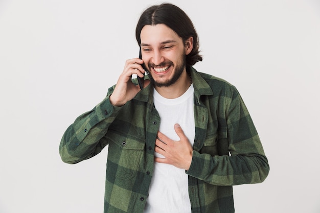 Portrait of an excited young bearded man wearing casual clothes standing isolated over wall, talking on mobile phone