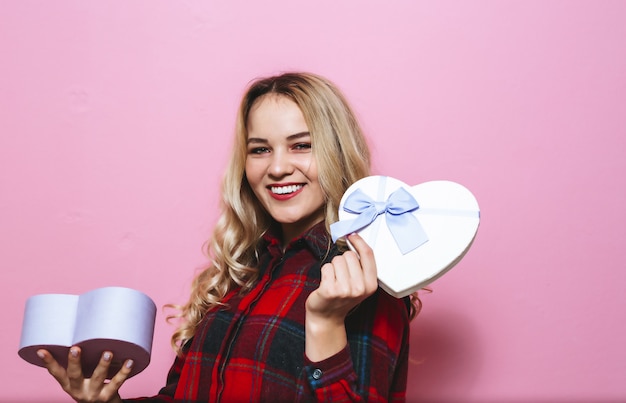 Портрет возбужденной молодой красивой блондинки, держащей подарок и счастливой над розовой стеной