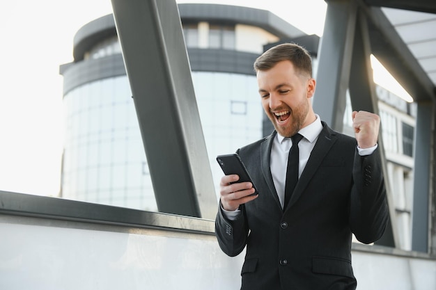 Portrait of an excited young businessman dressed in suit holding mobile phone celebrating