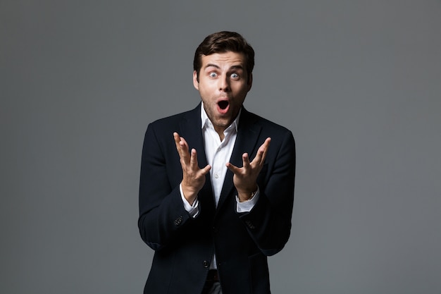 Portrait of an excited young businessman dressed in suit isolated over gray wall, celebrating success