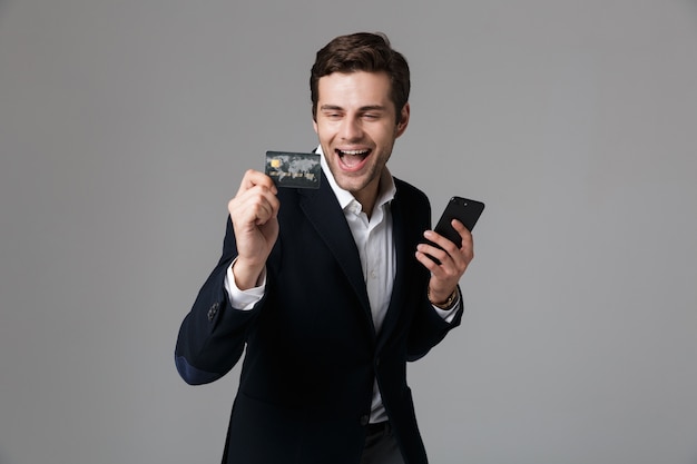 Portrait of an excited young businessman dressed in suit isolated over gray wall, using mobile phone, showing credit card