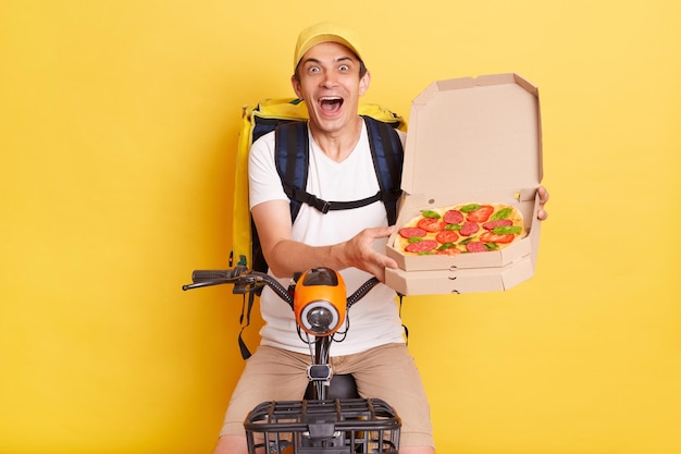 Portrait of excited young Caucasian courier man holding opened carton pizza box looking at camera with excitement screaming happily handing tasty order posing isolated over yellow background