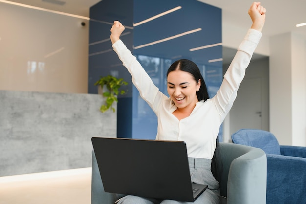 Ritratto di giovane donna caucasica eccitata che celebra il successo mentre si siede con il laptop a casa o in ufficio moderno felice freelance femminile che lavora nello spazio di coworking