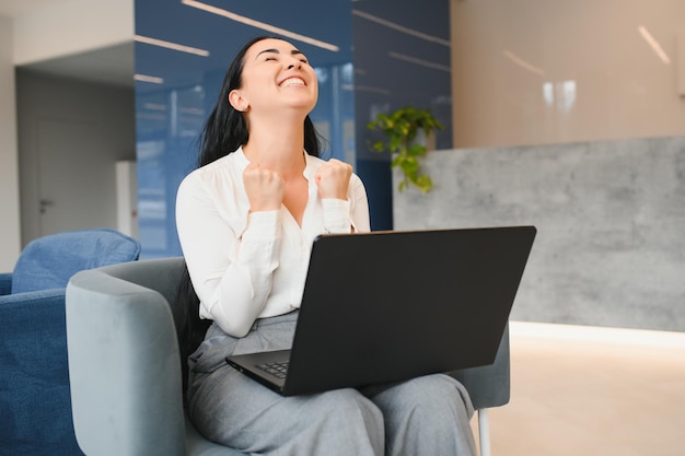 Portrait of excited young caucasian woman celebrating success while sitting with laptop at home or modern office Happy female freelancer working in coworking space