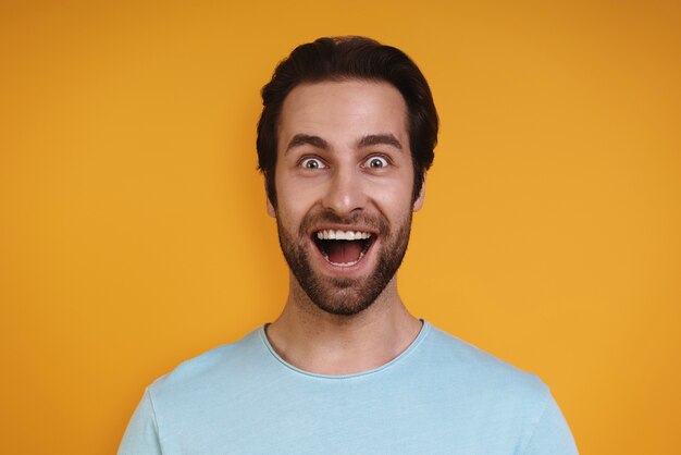 Portrait of excited young man in casual clothing looking at camera and smiling while standing agains...