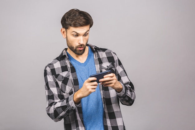 Portrait of an excited young man in casual playing games on mobile phone isolated over grey wall.