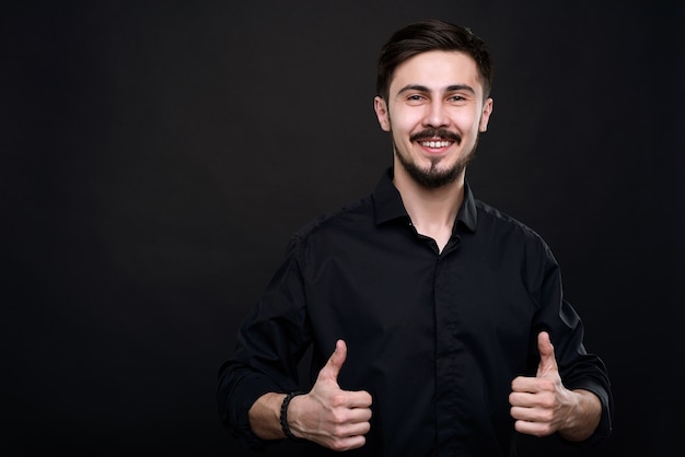 Portrait of excited young man standing against black wall and showing thumbs-up as symbol of quality