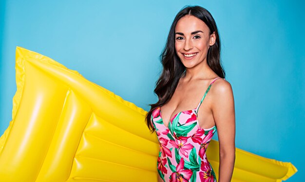 Portrait of an excited young smiling woman dressed in colorful swimsuit posing with yellow inflatable mattress isolated over blue background