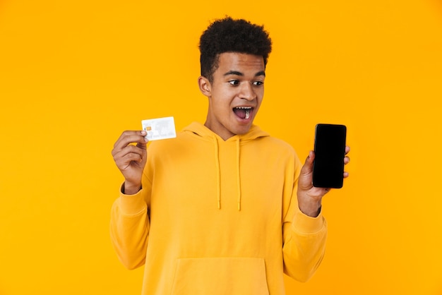 Portrait of an excited young teenager boy standing isolated over yellow wall, showing credit card and blank screen mobile phone