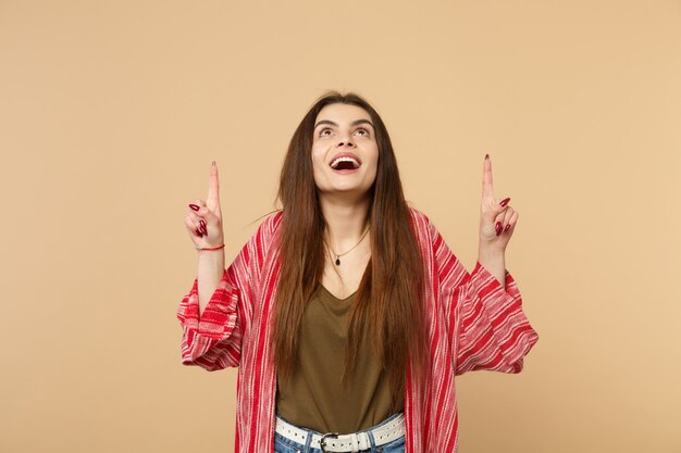 Portrait of excited young woman in casual clothes looking, pointing index fingers up isolated on pastel beige wall background in studio. People sincere emotions, lifestyle concept. Mock up copy space.