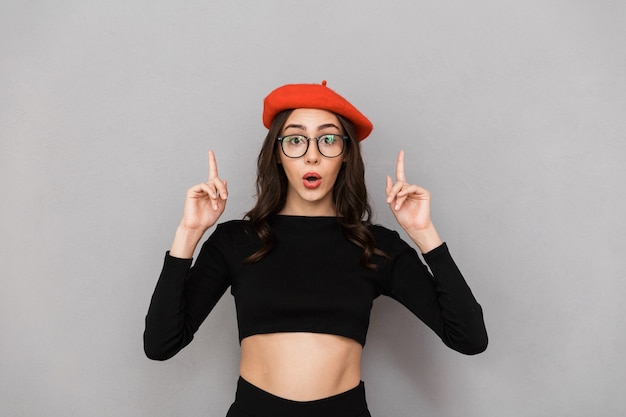 Portrait of an excited young woman dressed in red hat standing over gray background, pointing up