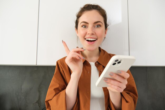 Photo portrait of excited young woman has an idea holding smartphone and pointing finger up standing in