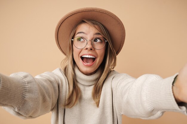 Portrait of excited young woman in hat smiling and taking selfie photo 