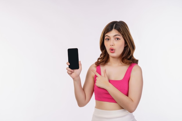 Portrait of excited young woman holding mobile phone mock up and pointing finger at smartphone isolated over white background