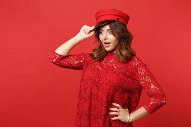 Portrait of excited young woman in lace dress holding cap, keeping mouth open looking aside isolated on bright red background in studio. People sincere emotions, lifestyle concept. Mock up copy space.