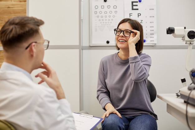 Portrait of excited young woman putting on new glasses and smiling happily in ophthalmology clinic