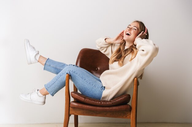 Portrait of excited young woman smiling and using headphones while sitting on armchair isolated on white