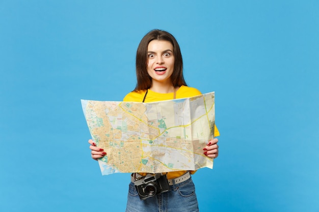 Portrait of excited young woman in vivid casual clothes with retro vintage photo camera holding paper city map isolated on blue wall background in studio. People lifestyle concept. Mock up copy space.