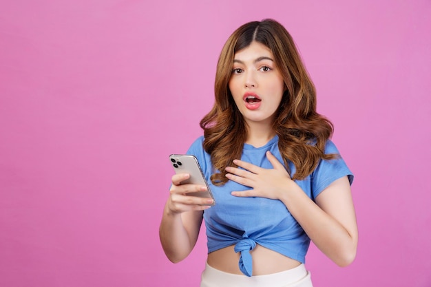 분홍색 배경 위에 격리된 스마트폰을 사용하여 캐주얼한 티셔츠를 입은 흥분한 젊은 여성의 초상화