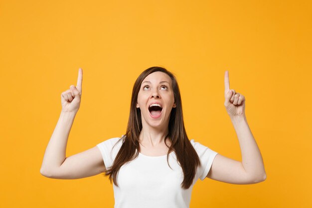 Portrait of excited young woman in white casual clothes keeping mouth open, pointing index fingers up isolated on yellow orange wall background in studio. People lifestyle concept. Mock up copy space.