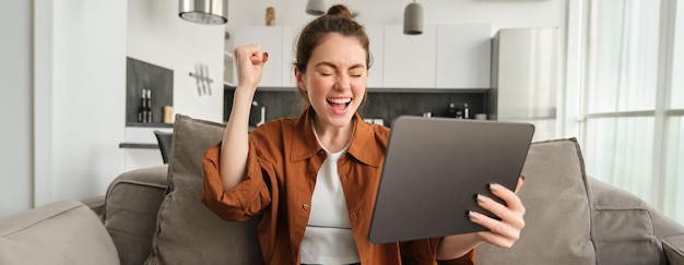 デジタルタブレットを持った興奮した若い女性の肖像画 ⁇ ソファに座って笑い ⁇ 笑顔で勝っています ⁇ 