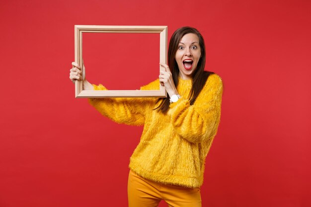 Portrait of excited young woman in yellow fur sweater keeping mouth wide open, holding picture frame isolated on bright red background. People sincere emotions, lifestyle concept. Mock up copy space.