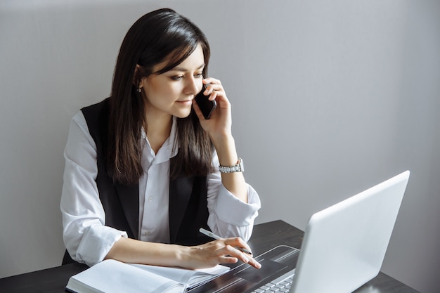 Портрет исполнительной молодой женщины, сидел на столе и работает на ноутбуке во время звонка