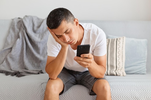 Portrait of exhausted sleepy bored man wearing casual style white t shirt sitting on cough keeps eyes closed having nap holding cell phone in hands looks tired