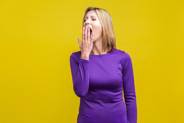 Portrait of exhausted sleepy woman in elegant tight purple dress yawning and covering mouth with hand feeling bored and tired lack of energy indoor studio shot isolated on yellow background