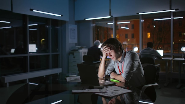Портрет измученного напряженного бизнесмена, работающего на ноутбуке ночью в темном офисе