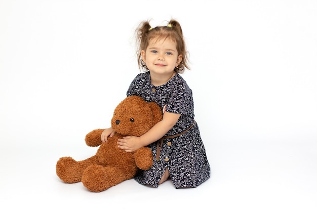 Portrait of expressive little girl hugging huge plush bear, indoor shot on white room. Little girl playing with teddy bear