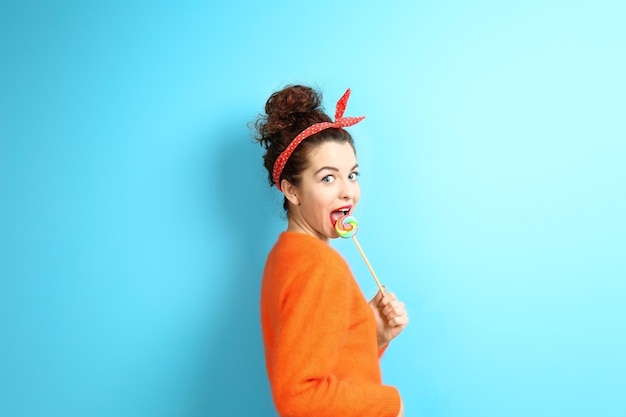 Portrait of expressive young model in orange sweater with lollipop on blue background