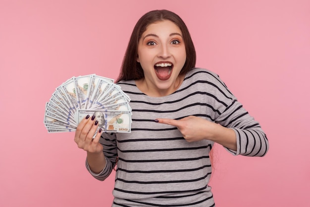 Portrait of extreme overjoyed delighted young woman in striped sweatshirt screaming in excitement and pointing dollar bills shocked by lottery win big jackpot isolated studio shot pink background