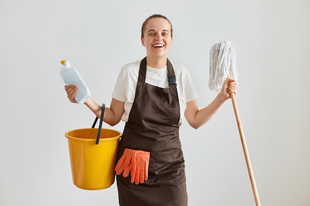 Портрет чрезвычайно счастливой кавказской женщины в белой футболке и фартуке, позирующей с чистящим оборудованием, держащей швабру и моющее средство, смотрящей на камеру, домохозяйка счастлива делать работу по дому