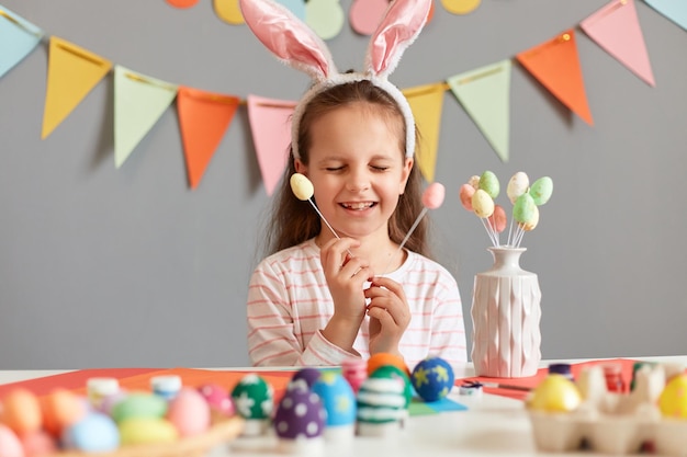 Portrait of extremely happy little girl holding multicolored painted dyed easter eggs on sticks keeps eyes closed smiling happily wearing bunny ears